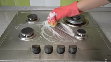 Eldivenli ev hanımı mutfaktaki gaz sobasını bez ve köpükle yıkıyor.