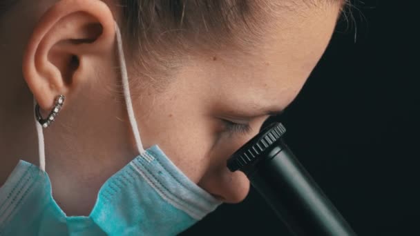 Chica científica mira en la lente de un microscopio en un primer plano de máscara — Vídeo de stock