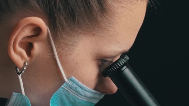 Chica científica mira en la lente de un microscopio en un primer plano de máscara — Vídeo de stock