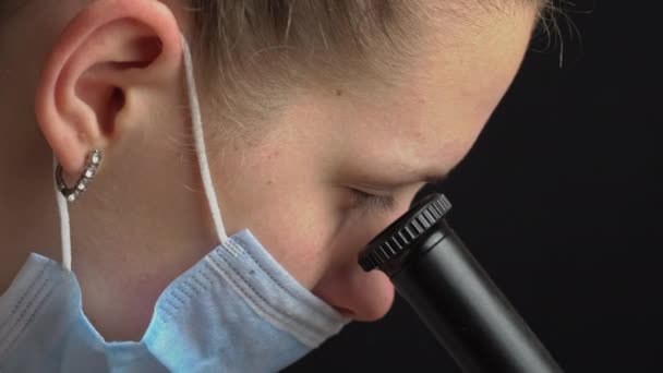 Ragazza scienziata guarda nella lente di un microscopio in una maschera Close-up — Video Stock