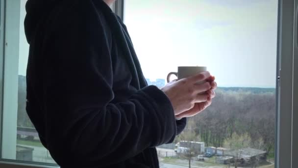 Mand drikker kaffe fra kop i sort badekåbe ved et åbent vindue om morgenen – Stock-video