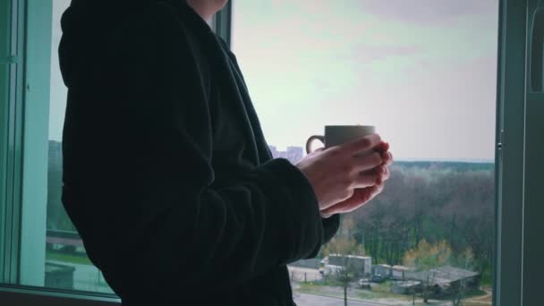 Человек пьет кофе из чашки в черном халате на открытом окне утром — стоковое видео