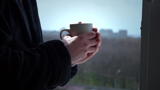 Muž pije kávu z poháru v černém županu v otevřeném okně v dopoledních hodinách — Stock video