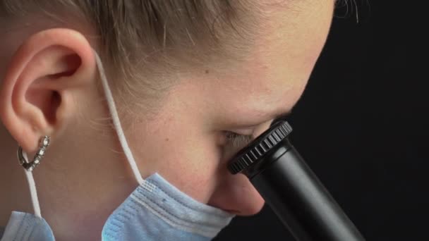 Chica científica mira en la lente de un microscopio en un primer plano de máscara — Vídeo de stock