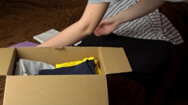 Genç bir kız, içinde eşyalar olan bir paket topluyor ve elbiseleri inceliyor. Kadın güzel şeyleri kahverengi bir kutuya koyar. Nesnelerin internet ve posta yoluyla teslimatı.