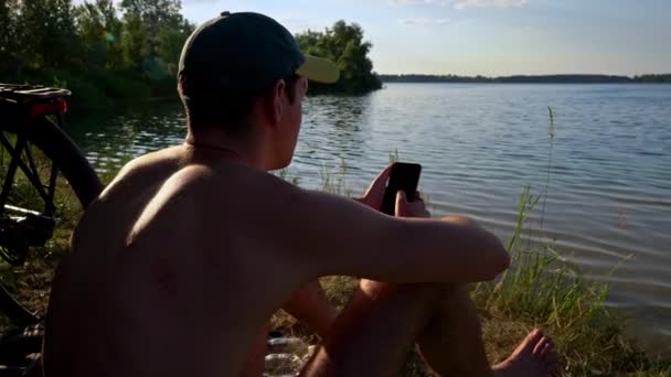 Joven Con Gorra Sienta Cerca Del Río Teléfono Inteligente Naturaleza — Vídeos de Stock