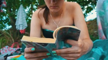 Genç kız sahilde yavaş çekim mayosuyla kitap okuyor. Havlu giymiş bir kadın yatak örtüsüne oturur ve ilginç bir kitap okur. Nehir kenarındaki doğada rahatla..