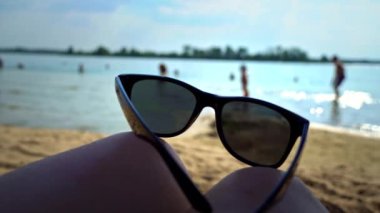 Sahilin arka planındaki bir kızın bacaklarında plaj manzarası gözlükleri. Deniz suyunun arka planında güneş gözlüğü.