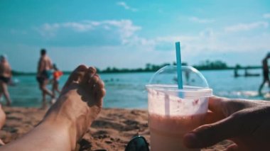 Kadın bacaklarının ve plajın arka planında eller milkshake alır. Deniz teması, rahatlama. Bir kızın bacakları bir bardağın yanında, denizin arka planında pipetle..