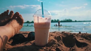 Milkshake ve plajın arka planında kadın bacakları. Deniz teması, rahatlama. Bir kızın bacakları bir bardağın yanında, denizin arka planında pipetle..