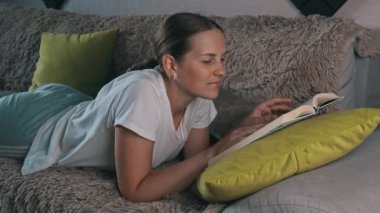 Girl is reading a book at home with her domestic gray cat. Happy lady in casual wear smiling and relaxing at home with her pet. Home leisure concept.