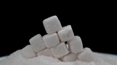 White sugar cubes fall on granulated sugar on a black background. The sugar spreads evenly. Benefits or harms of sugar consumption for health.