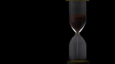Sand in the hourglass crumbles in the dark. The countdown hourglass is pouring on a black background. Sand falls into a glass flask. Sand is poured onto the flask of the hourglass.