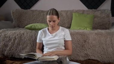 Girl is reading a book at home with her domestic gray cat. Happy lady in casual wear smiling and relaxing at home with her pet. Home leisure concept.