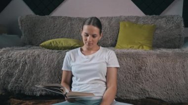 Girl is reading a book at home with her domestic gray cat. Happy lady in casual wear smiling and relaxing at home with her pet. Home leisure concept.