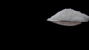 Sugar cubes fall into a teaspoon on a black background in slow motion. White sugar cubes fall on granulated sugar. The sugar spreads evenly. Benefits or harms of consumption for health.