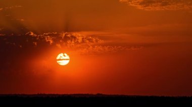 Time lapse bright orange sunset sun sets behind the clouds. The end of the day, the sun goes below the horizon. Big red hot sun in warm air distortion above the horizon.