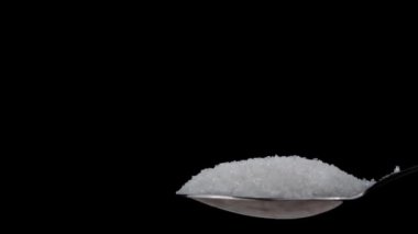 Sugar cubes fall into a teaspoon on a black background in slow motion. White sugar cubes fall on granulated sugar. The sugar spreads evenly. Benefits or harms of consumption for health.