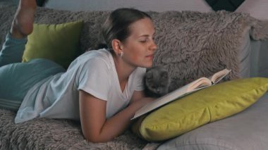 Girl is reading a book at home with her domestic gray cat. Happy lady in casual wear smiling and relaxing at home with her pet. Home leisure concept.