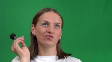 Güzel kız yüzünü yeşil bir arka planda fırçayla boyuyor. Krom adasında, yeşil ekranda izole edilmiş, güzellik dünyasındaki genç bir kadın. Kız yüzünü boyuyor..