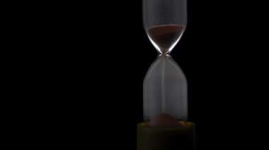 Sand in the hourglass crumbles in the dark. The countdown hourglass is pouring on a black background. Sand falls into a glass flask. Sand is poured onto the flask of the hourglass.