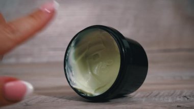 Masanın üzerinde bir kavanoz krema var ve bir kadının parmağı alıyor. Kadın yüzünü ve vücudunu nemlendiriyor. Kadınların güzelliği ve cilt bakımı. Güzel kadın tırnakları.