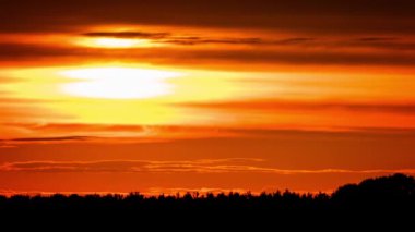 Time lapse bright orange sunset sun sets behind the clouds. The end of the day, the sun goes below the horizon. Big red hot sun in warm air distortion above the horizon.
