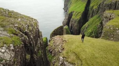 Dunnesdrangar, Faroe Adaları 'na bakan bir adamın 4K hava aracı görüntüsü. İnanılmaz doğa, Faroe Adaları 'ndaki büyük kayalıklar ve dağlar. Vagar Adası. Bir sürü uçan deniz kuşu. Yazın güneşli bir gün.