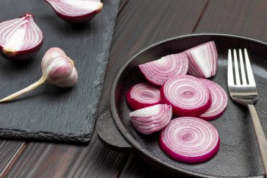 Kızartma tavasında dilimlenmiş mor soğan halkaları. Soğan yarımları ve bir baş sarımsak. Koyu ahşap arka plan. Üst görünüm