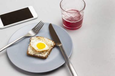 Çatal ve bıçak, rendelenmiş peynirli tost ve gri tabakta kaynamış bıldırcın yumurtası. Bardakta meyve suyu, masada cep telefonu. Gri arka plan. Uzayı kopyala Üst görünüm