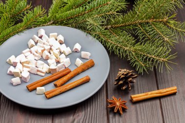 Marshmallows  and cinnamon sticks in ceramic plate. Star anise and pinecone on table. Pine branches. Top view. Dark wooden background.