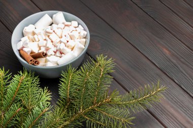 Dessert marshmallows  and cinnamon sticks in ceramic bowl. Pine branches. Copy space. Dark wooden background.