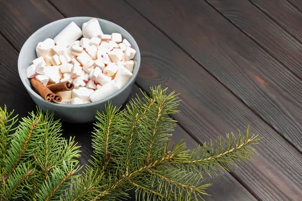 Dessert marshmallows  and cinnamon sticks in ceramic bowl. Pine branches. Copy space. Dark wooden background.