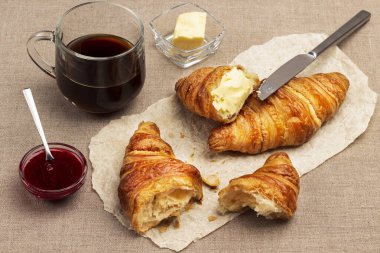 Kahvaltıda. Tereyağlı kruvasan. Kasede reçel, bir fincan kahve. Çuval bezleri masaya. Üst görünüm