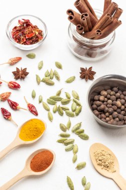 Metal kasede Allspice ve kavanozda tarçın çubukları. Ahşap kaşıklarda kırmızı biber, zerdeçal ve zencefil baharatları. Masada kakule tohumları ve kuru biber kabukları var. Üst Manzara. Beyaz arkaplan