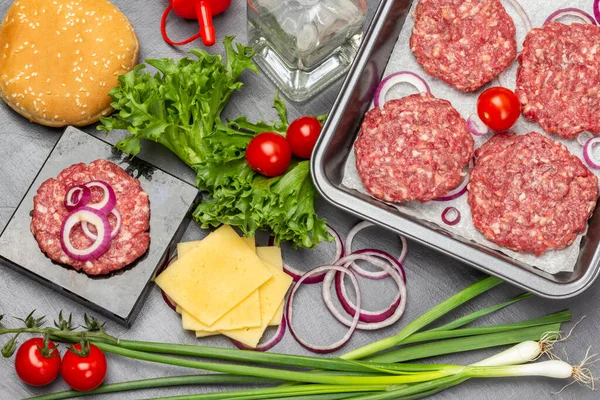 Yeşil soğan, peynir, su şişesi. Susamlı çörekler, yeşil salata ve domates. Kızartma tavasında çiğ sığır burger. Düz yatıyordu. Koyu arkaplan.