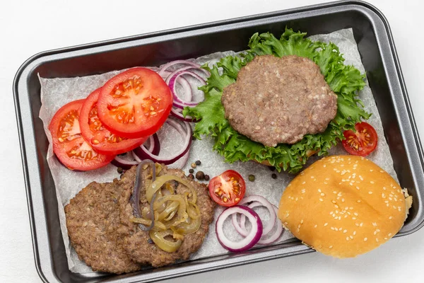 Fried burger bun on lettuce leaf, fried burger with cheese and fried onions, chopped tomatoes and onions on baking tray. Flat lay. White background