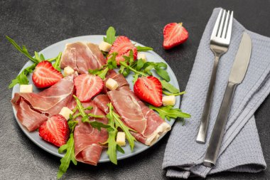 Çatal ve bıçak gri peçeteye. Peynirli İspanyol jamonu, gri tabakta çilek ve roka. Üst Manzara. Siyah arkaplan