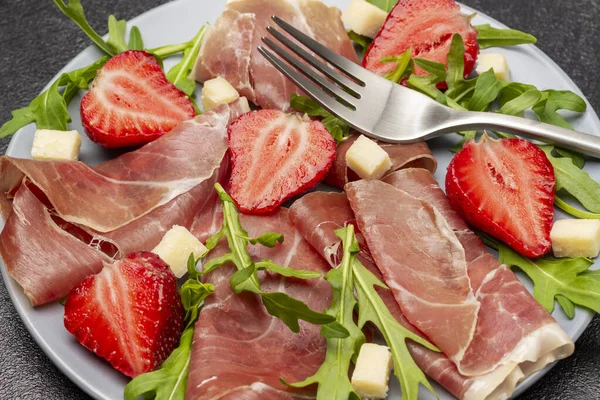 İspanyol jamonu, gri tabakta çilek ve roka. Kapatın. Üst Manzara. Siyah arkaplan