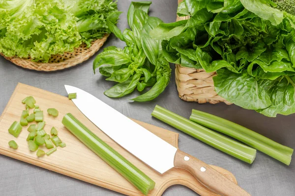 Knife and celery stalks on cutting board. Lettuce leaves in wicker basket and plate. Top view. Grey background.