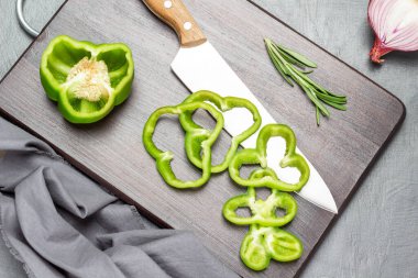 Kitchen knife and chopped green pepper on cutting board. Half an onion and grey napkin. Flat lay. Grey background.