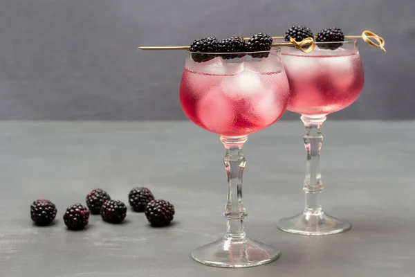 Two misted wine glasses with cooling drink and ice. Blackberry on skewer on wine glass. Blackberry on table. Flat lay. Grey background