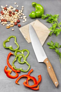 Chopped green and red pepper. Kitchen knife on paper, nuts and chickpeas, parsley sprigs on table. Flat lay. Grey background.