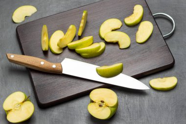 Two halves of green apple. Kitchen knife and apple slices on cutting board. Flat lay. Grey background.