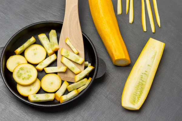 Sliced yellow zucchini and wooden spatula in frying pan. Whole yellow zucchini on table.  Flat lay. Grey background