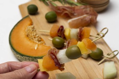 Elleri peynirli, zeytinli ve jambonlu şiş tutuyor. Kavun, jambon ve zeytin. Kapatın. Beyaz arkaplan.