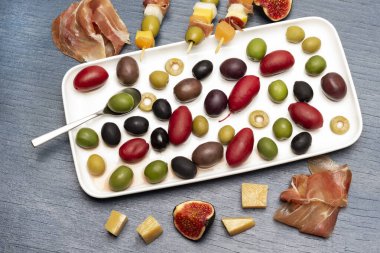 Jamon, peynir, incir ve peynirli şiş, zeytin ve reçel. Tabakta çeşitli zeytinler var. Düz yatıyordu. Mavi arkaplan.