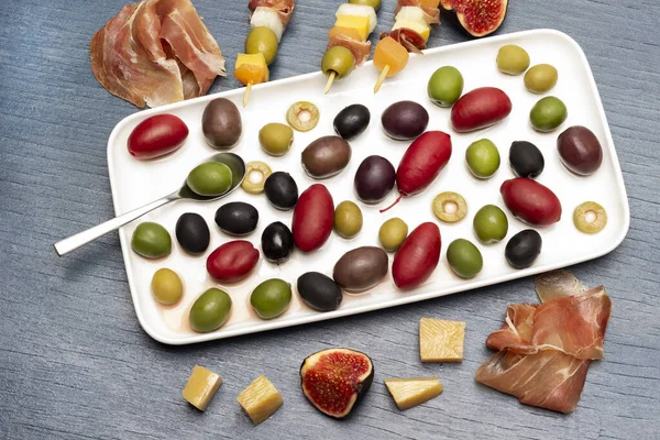 Jamon, peynir, incir ve peynirli şiş, zeytin ve reçel. Tabakta çeşitli zeytinler var. Düz yatıyordu. Mavi arkaplan.