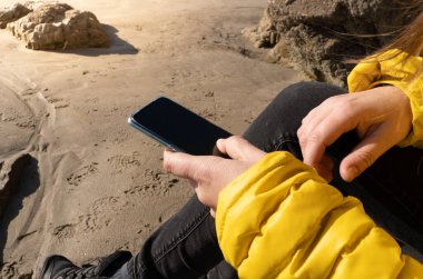 Sahilde elinde cep telefonu olan sarı ceketli bir kadın.