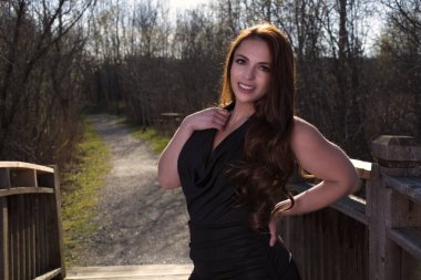 Gülümseyen genç bir kadın, bahar parkında poz veriyor. Siyah bir elbise giyiyor.